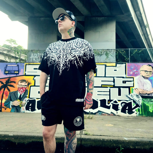 Person wearing a black and white patterned t-shirt and black shorts with tattoos, standing in front of graffiti art. RTLA Apparel