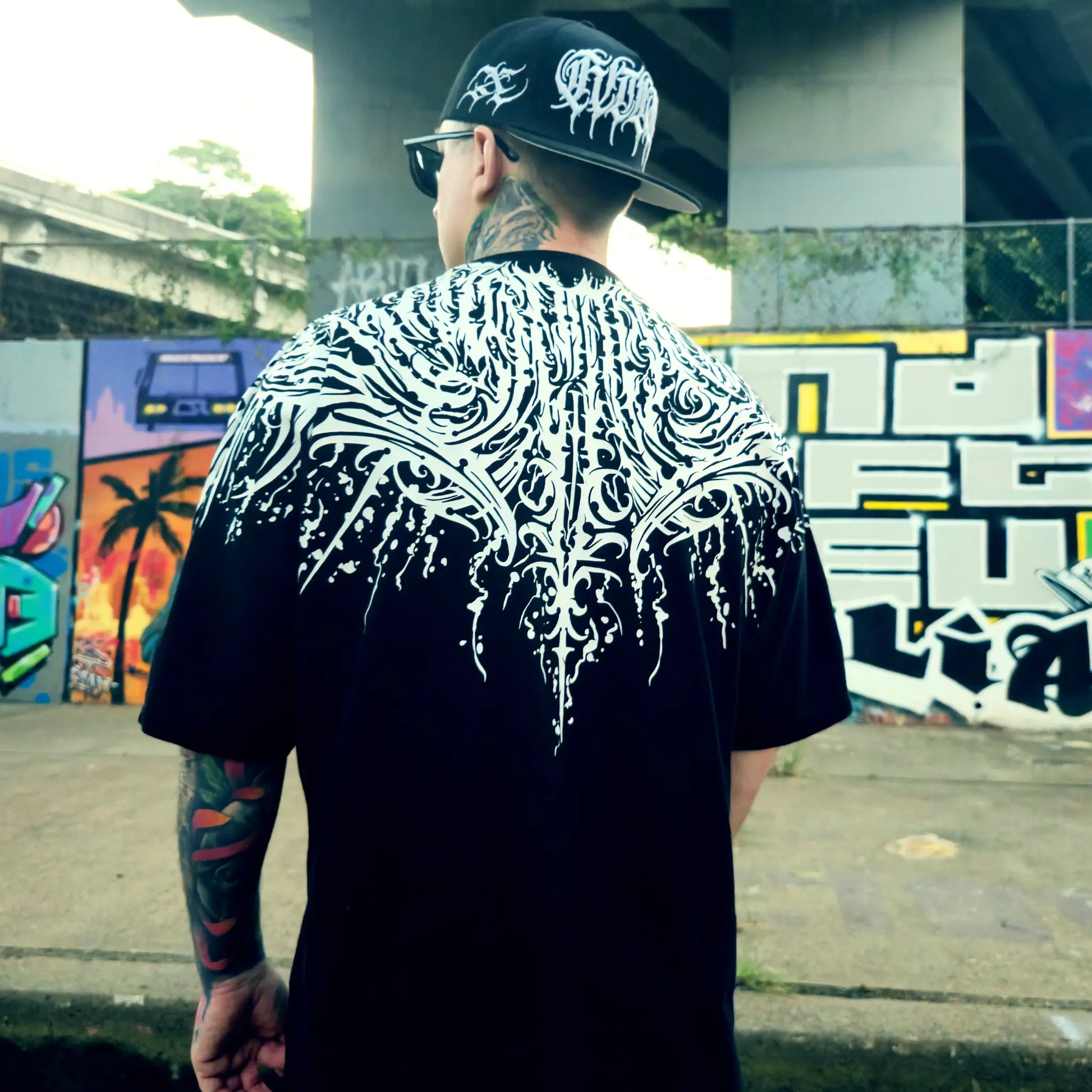 Person wearing a black t-shirt with white intricate design, standing in an urban setting with graffiti. RTLA Apparel