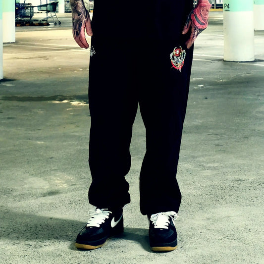 A person wearing black sweatpants with a red and white graphic patch on the left thigh, paired with black sneakers featuring white laces and white Nike swooshes, standing in a concrete parking structure.
