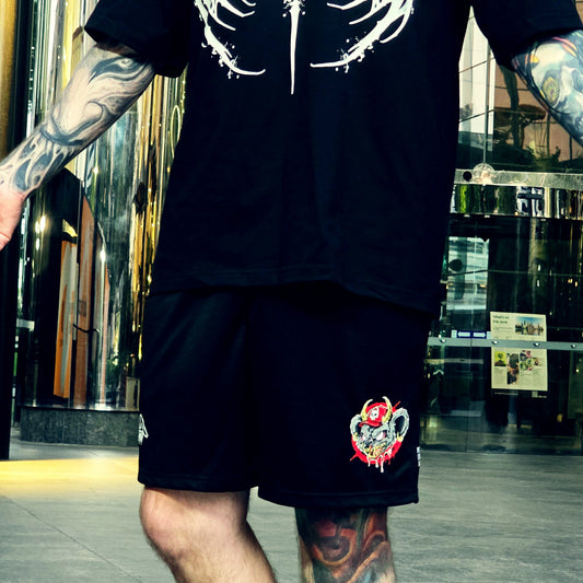A person wearing a black t-shirt and black athletic shorts with graphic prints, standing in front of a modern glass building.