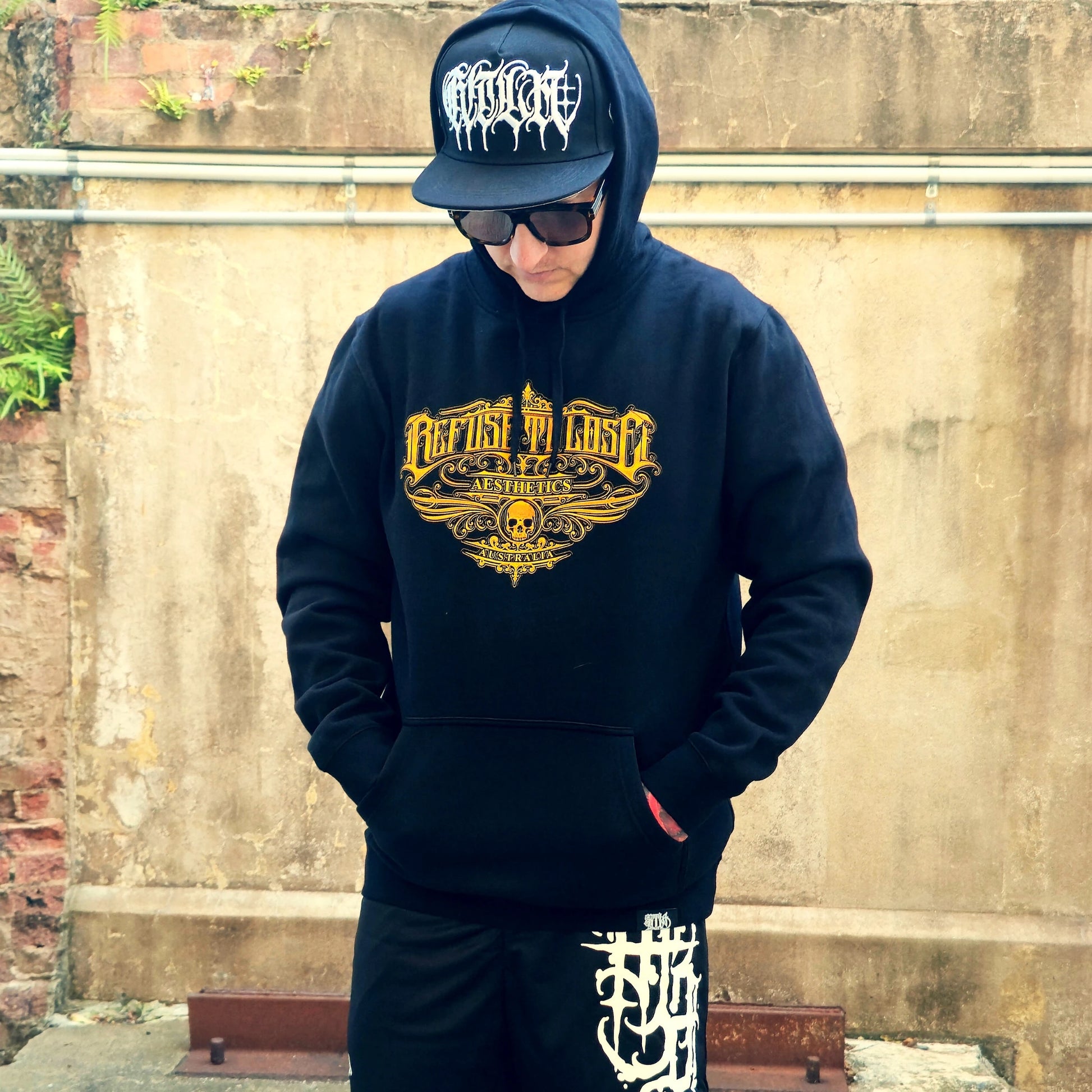 Person wearing a black hoodie with gold graphic design and a blue cap, standing against a textured wall.