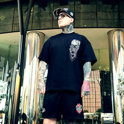 Person wearing a black t-shirt with a graphic design and black shorts with a logo, standing in front of a modern building.