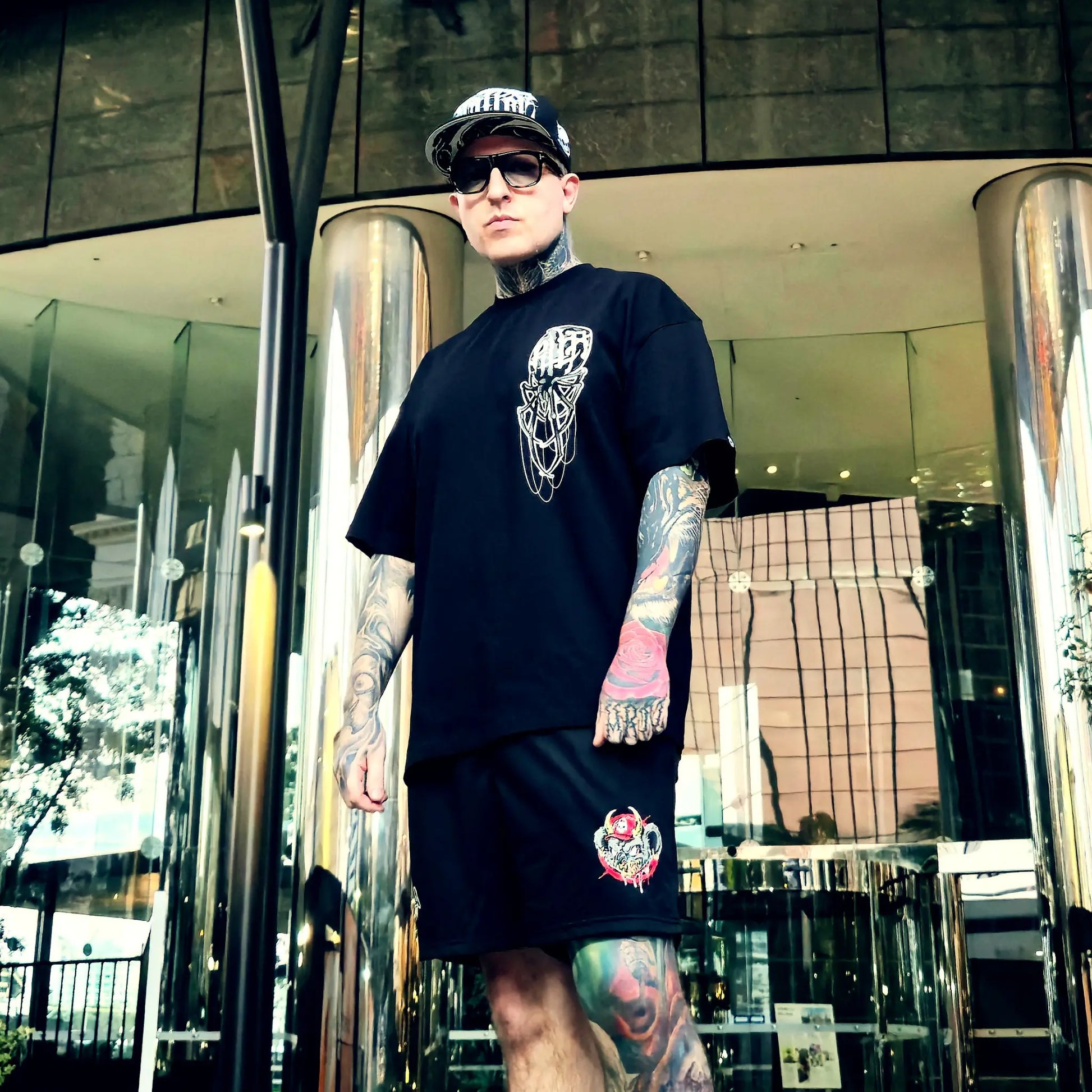 Man wearing a black t-shirt and shorts with tattoos standing in front of a modern building.