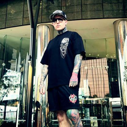 Man wearing a black t-shirt and shorts with tattoos standing in front of a modern building.