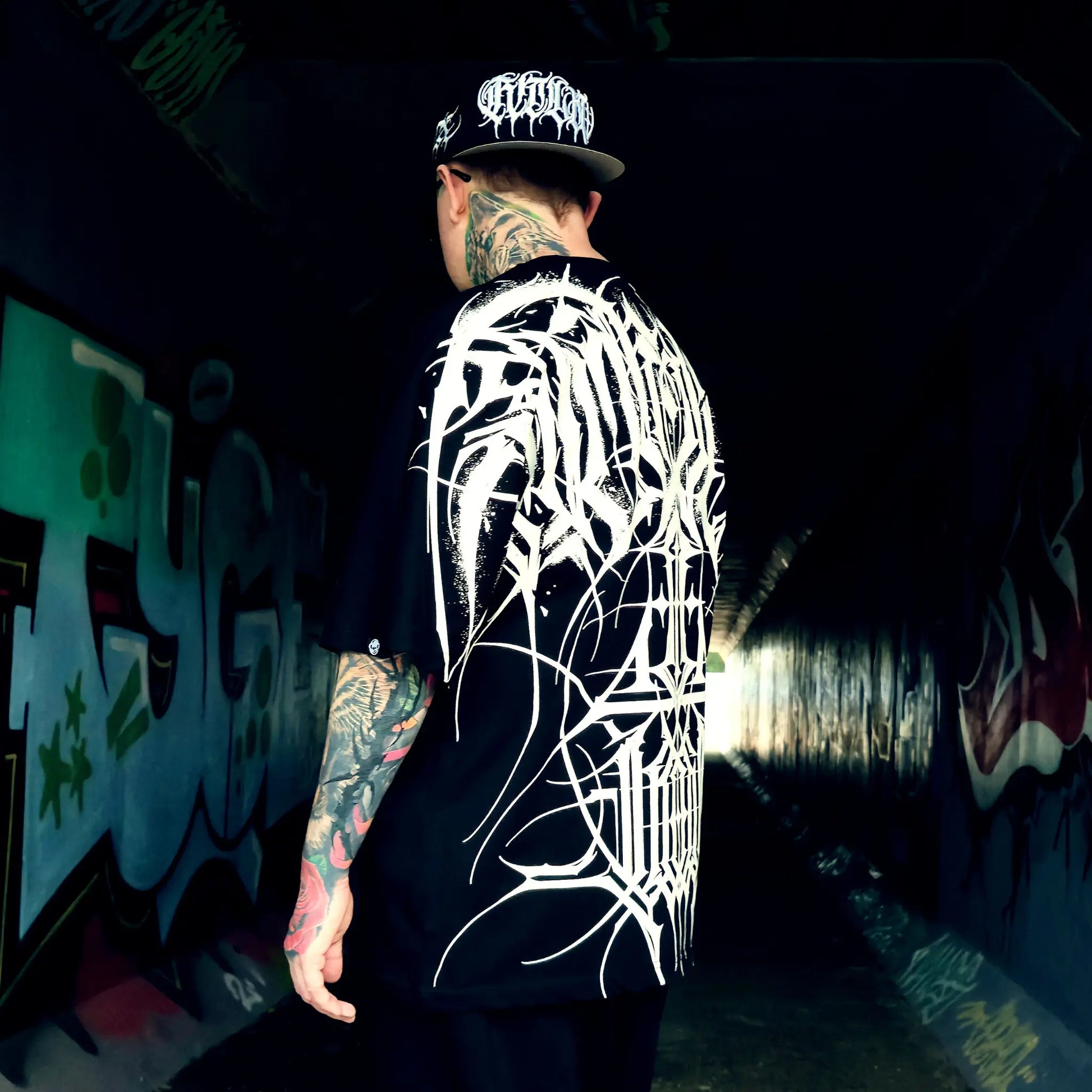 Person wearing a black t-shirt with white graphic design and cap, standing in a graffiti-covered tunnel.