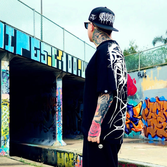 Person wearing a black cap and black shirt with white designs, standing in front of colorful graffiti.
