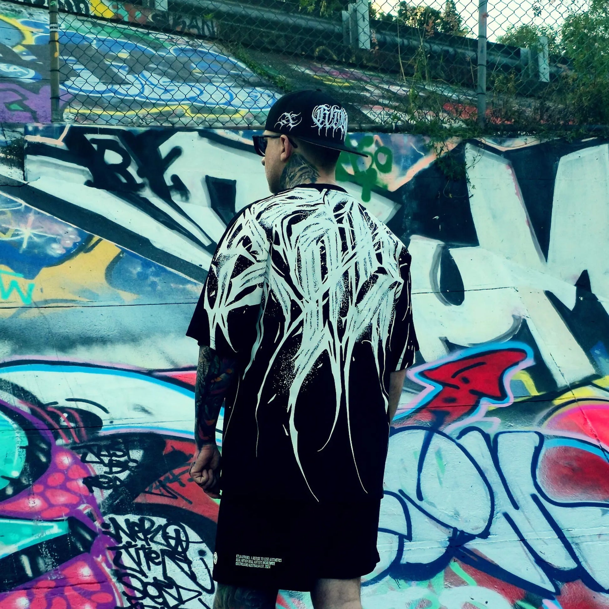 Person wearing a black and white outfit with a cap, standing in front of a colorful graffiti wall.