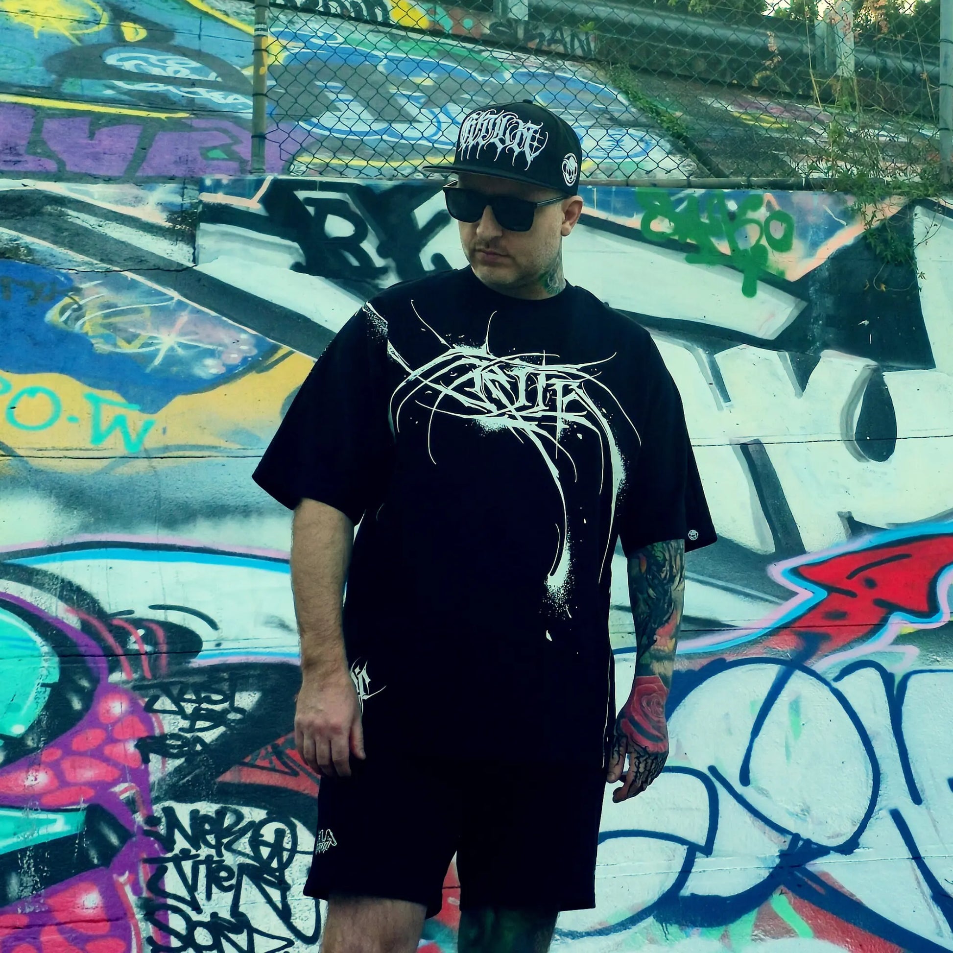 Person wearing a black t-shirt with white graphics and a black cap, standing in front of a colorful graffiti wall.