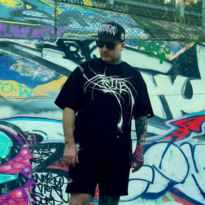 Person wearing a black t-shirt with white graphics and a black cap, standing in front of a colorful graffiti wall.