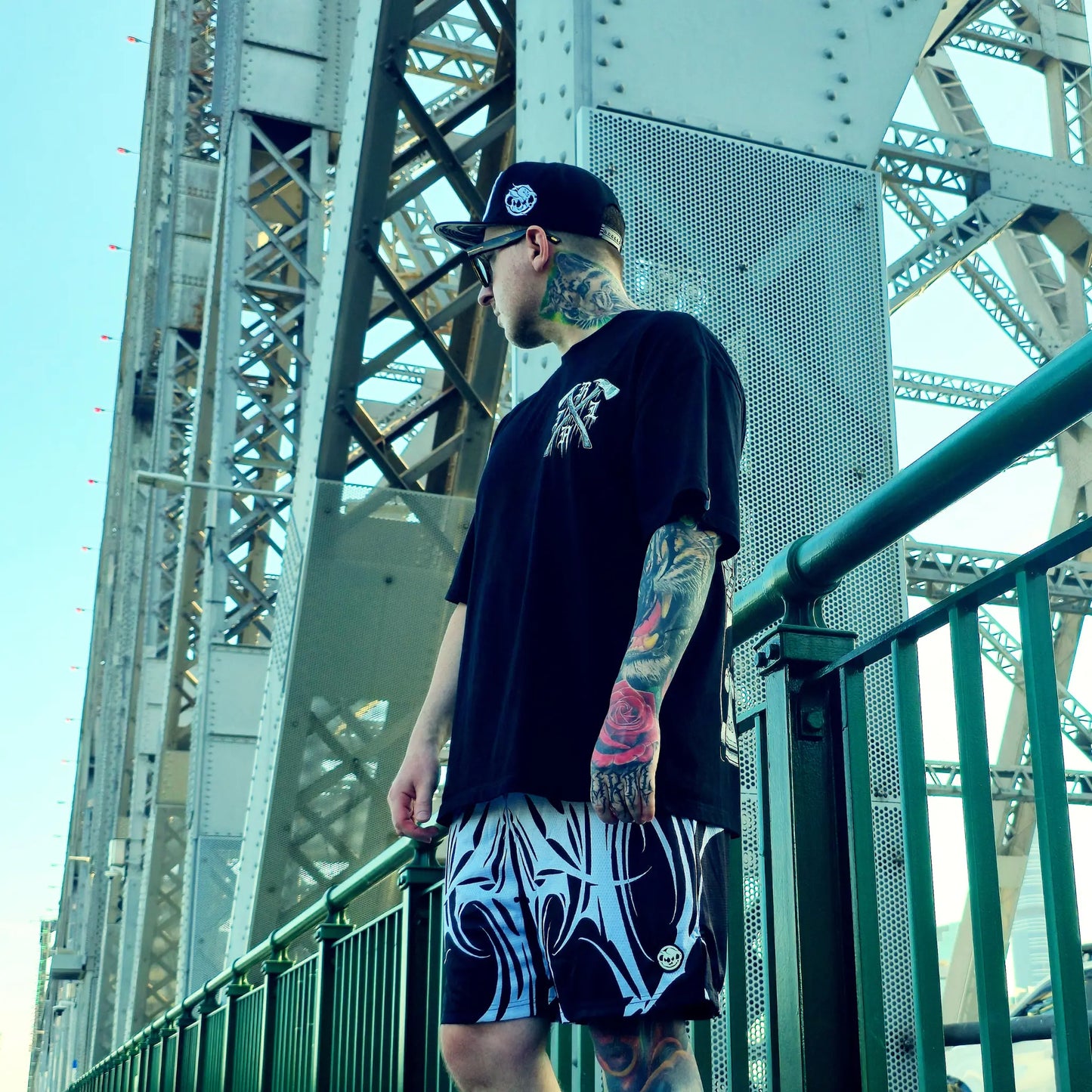 Man with tattoos wearing a black t-shirt and patterned shorts standing on a bridge.