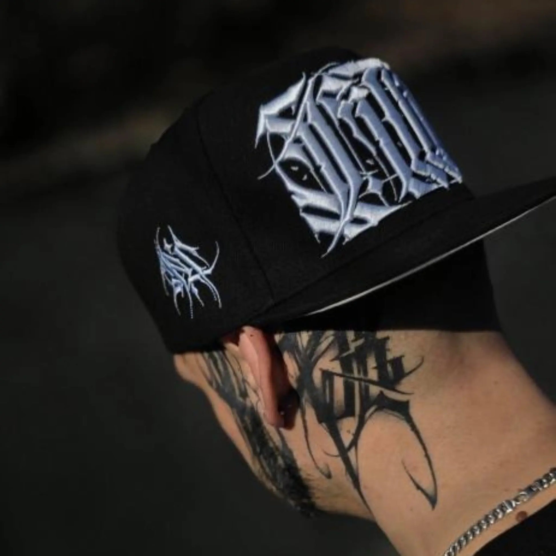 Black cap with white embroidered design worn by a person with tattoos on a dark background