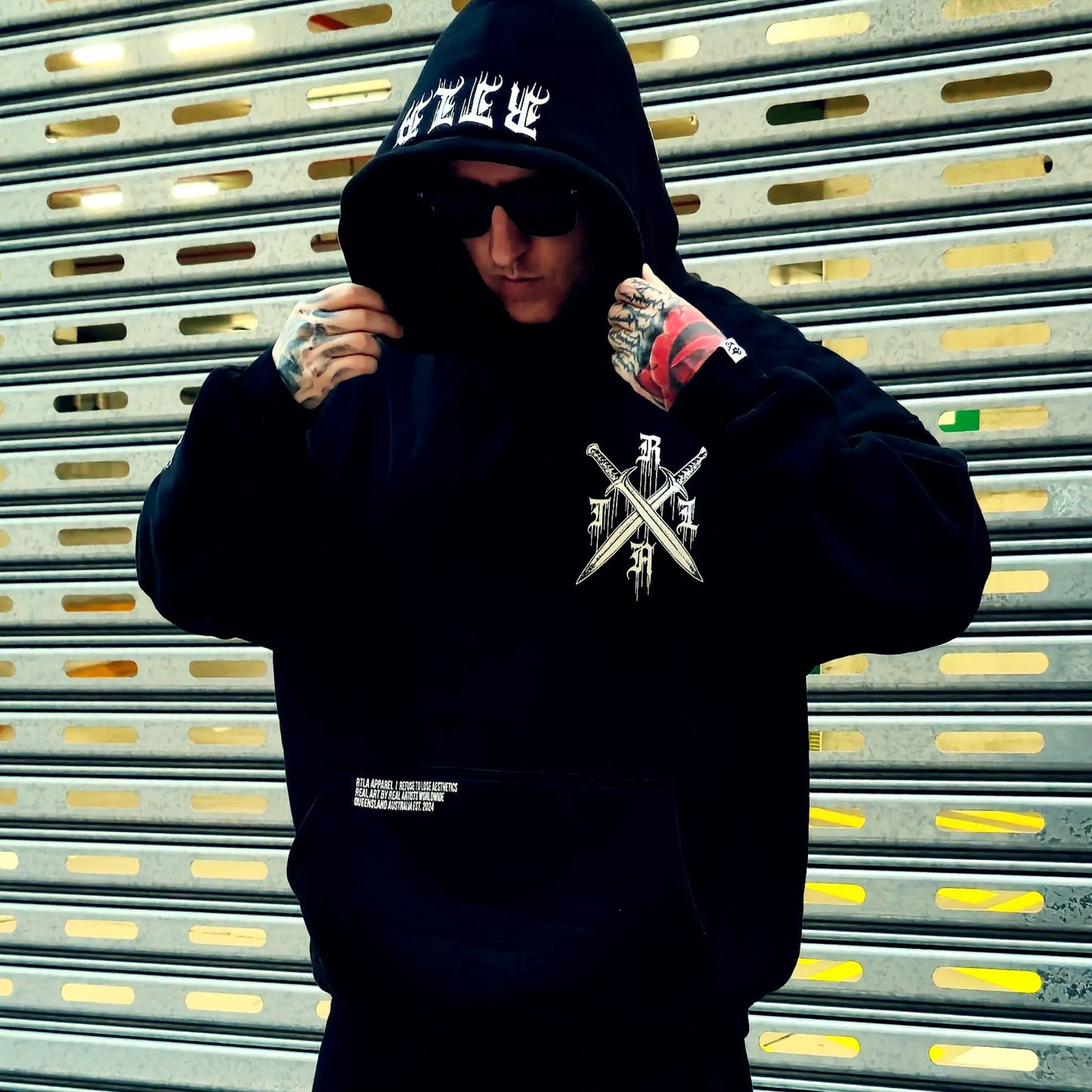 Person wearing a black hoodie with a logo in front of a metallic shutter.