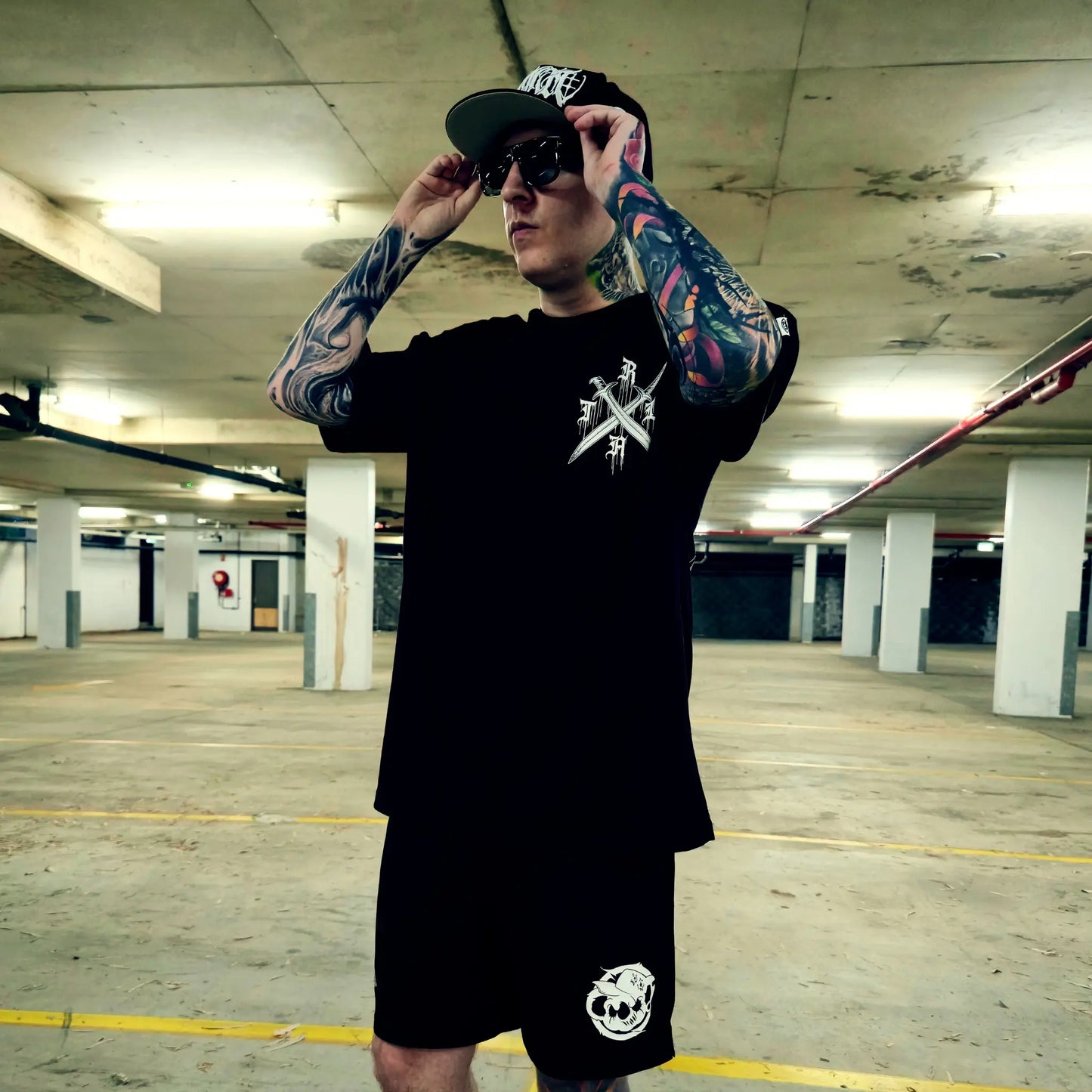 Man with tattoos wearing a black t-shirt and shorts in an underground parking lot.