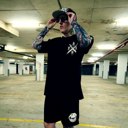 Man with tattoos wearing a black t-shirt and shorts in an underground parking lot.