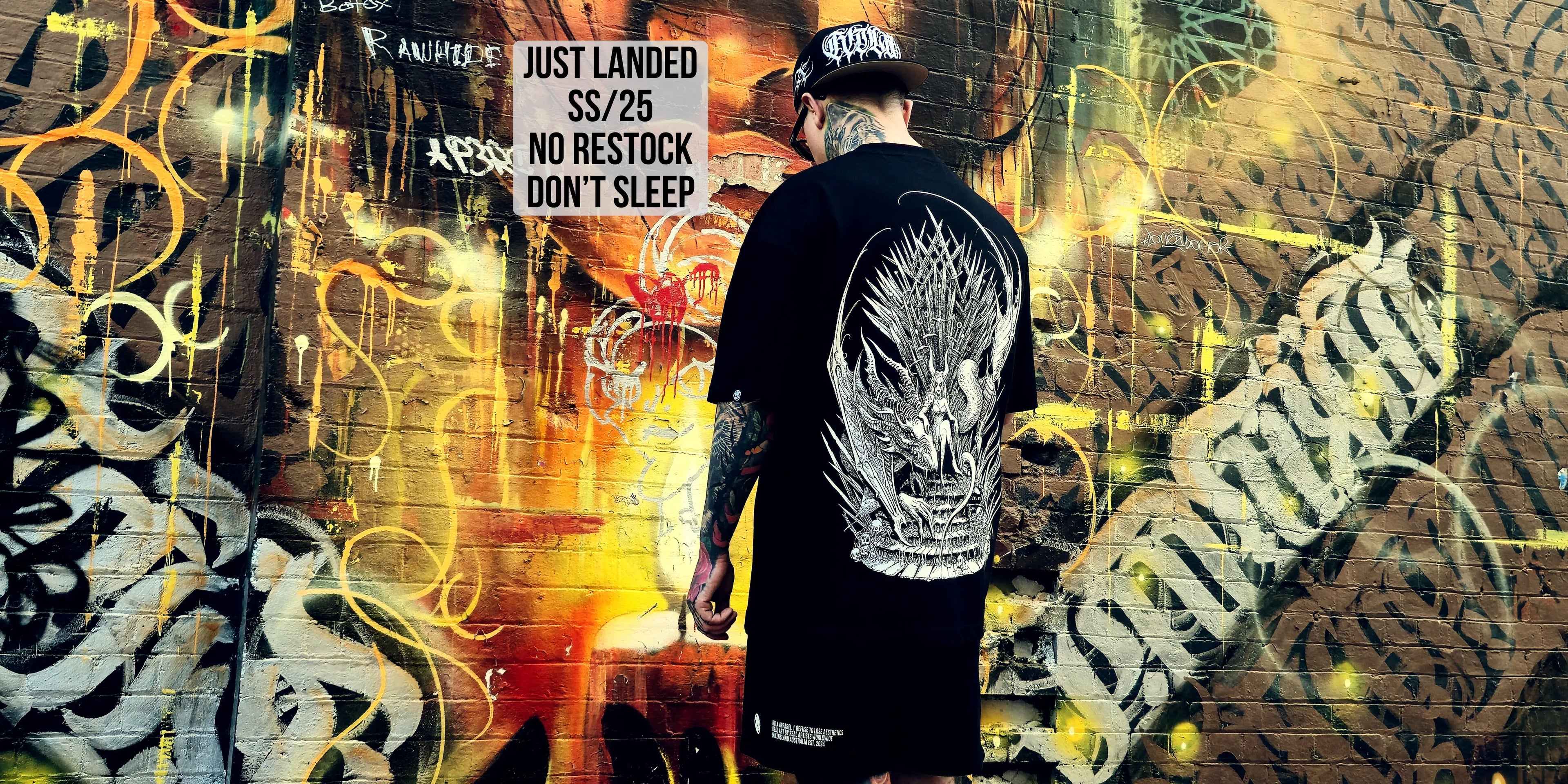Person wearing a black t-shirt with a white graphic design against a graffiti-covered wall.