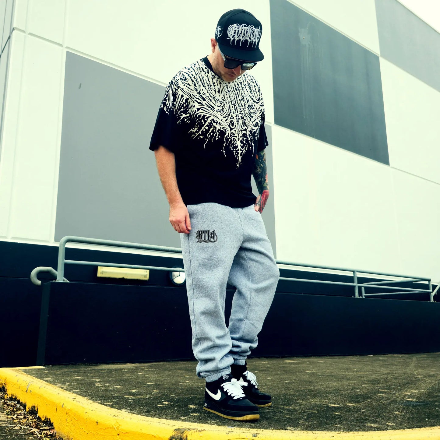 Person wearing a black t-shirt with white graphics, gray sweatpants, and black sneakers in an urban setting.