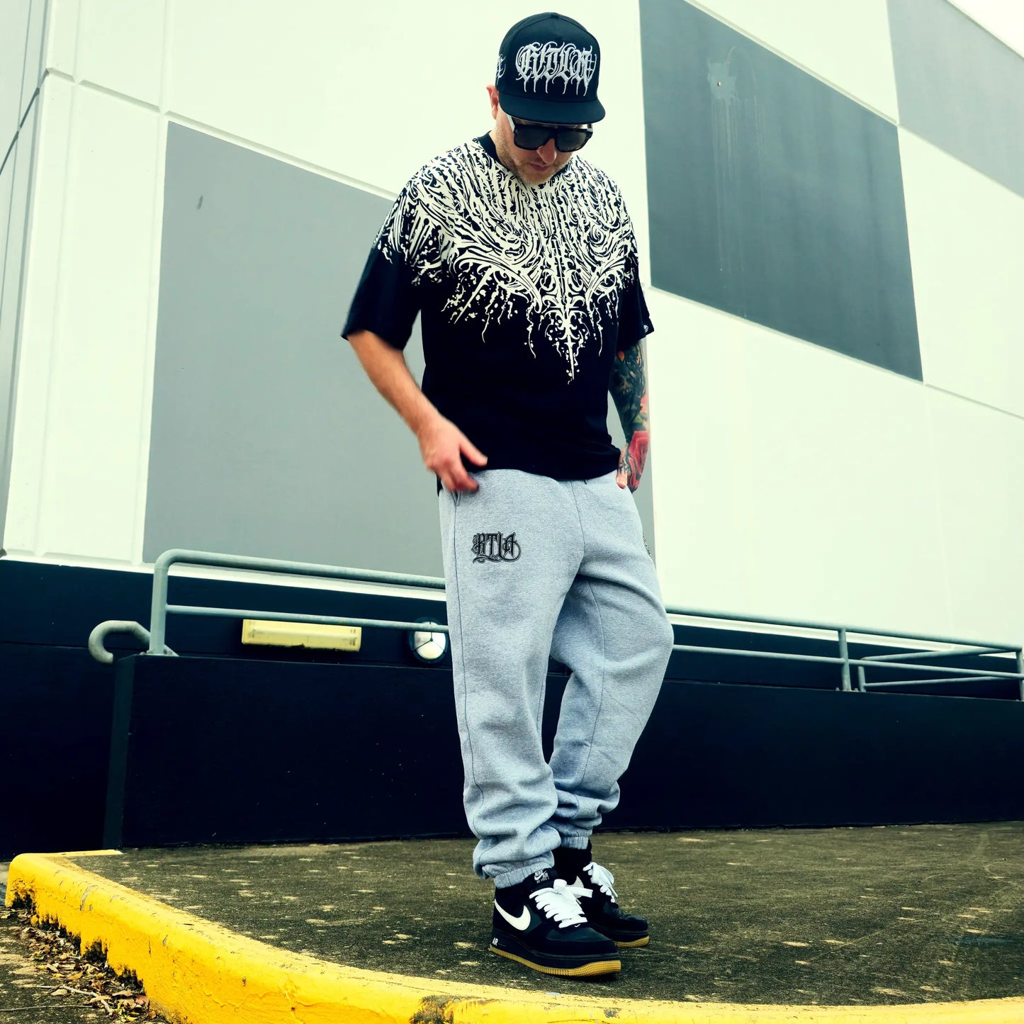 Person wearing a black and white patterned shirt, gray sweatpants, and black sneakers with a cap, standing on a yellow curb.