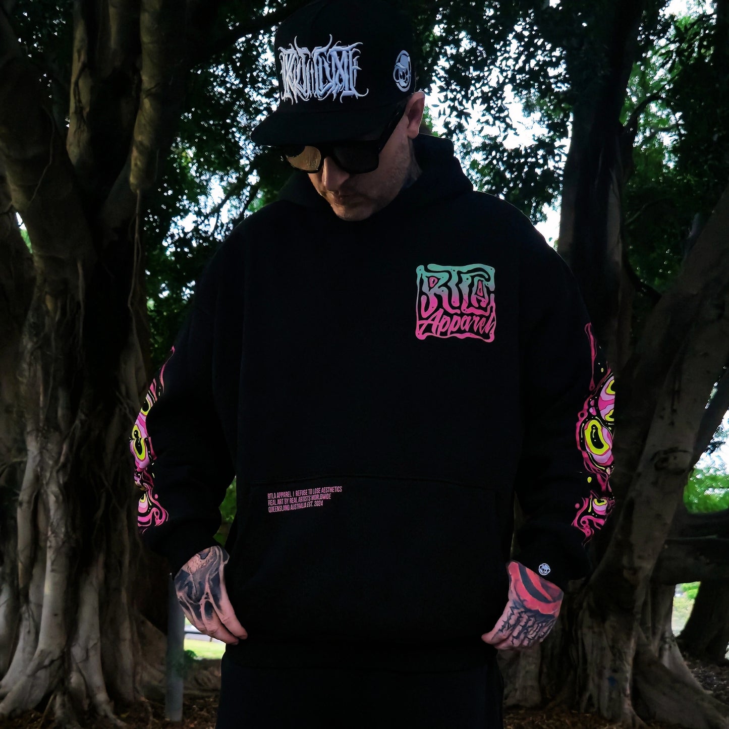 Person wearing a black hoodie with colorful graphics and text, standing in a forest setting.