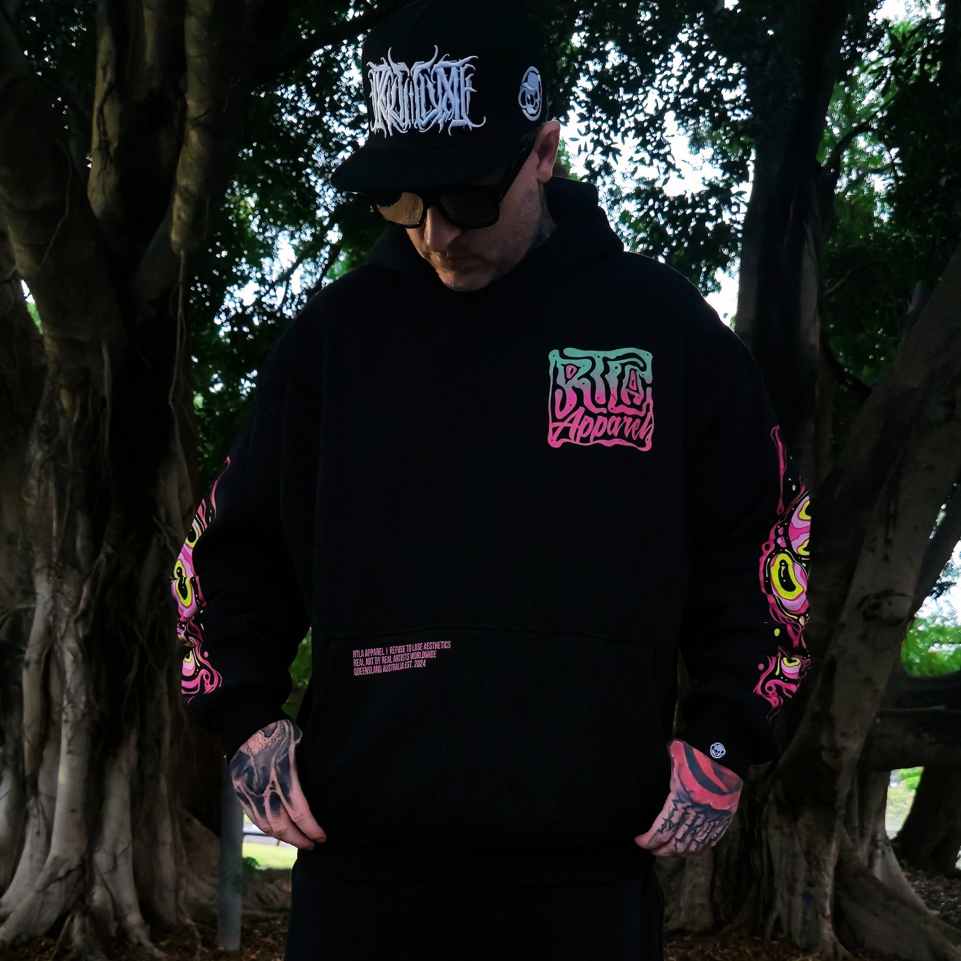 Person wearing a black hoodie with colorful graphics and text, standing in a forest setting.