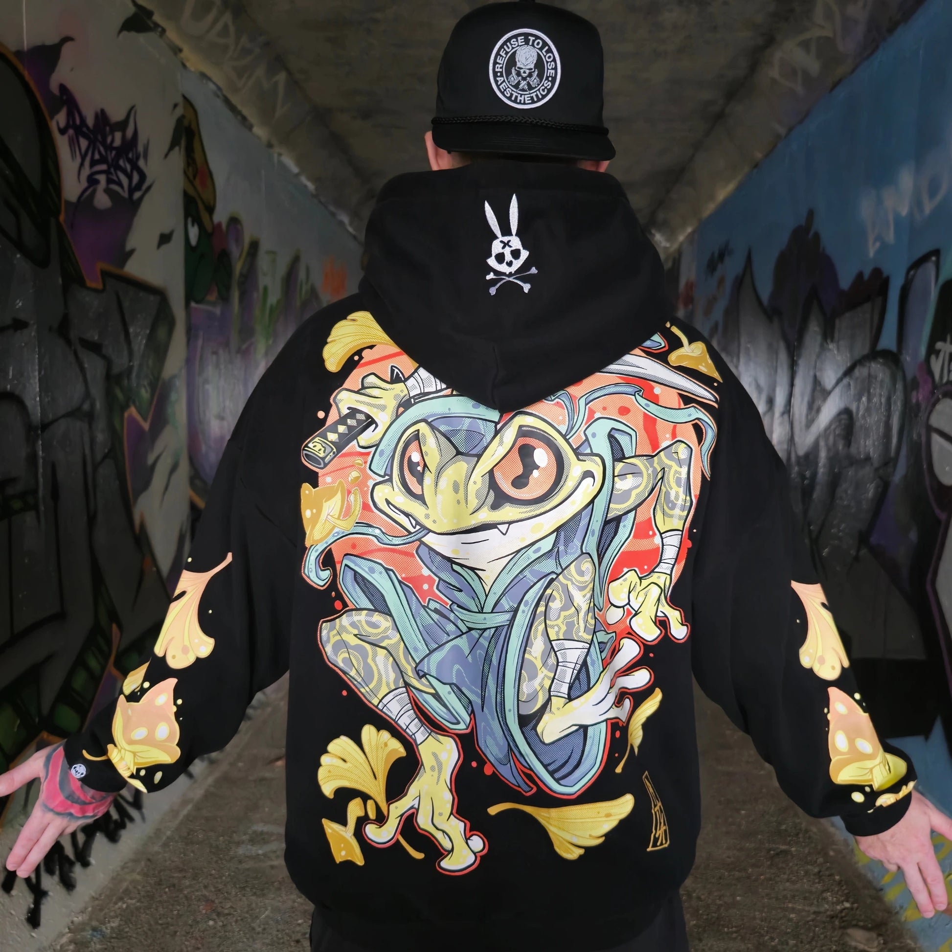 A person wearing a black oversized hoodie with a colourful character design on the back, standing in a graffiti-covered hallway. RTLA Apparel
