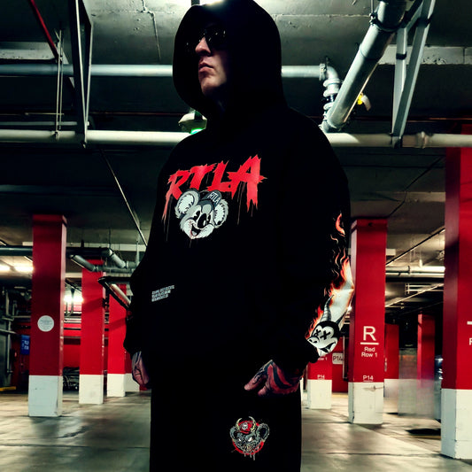 A person wearing a black hoodie with a red and white graphic print stands in a parking garage. The hoodie features a large red and white graphic on the chest, a flame design on the sleeve, and a small graphic near the lower front pocket.