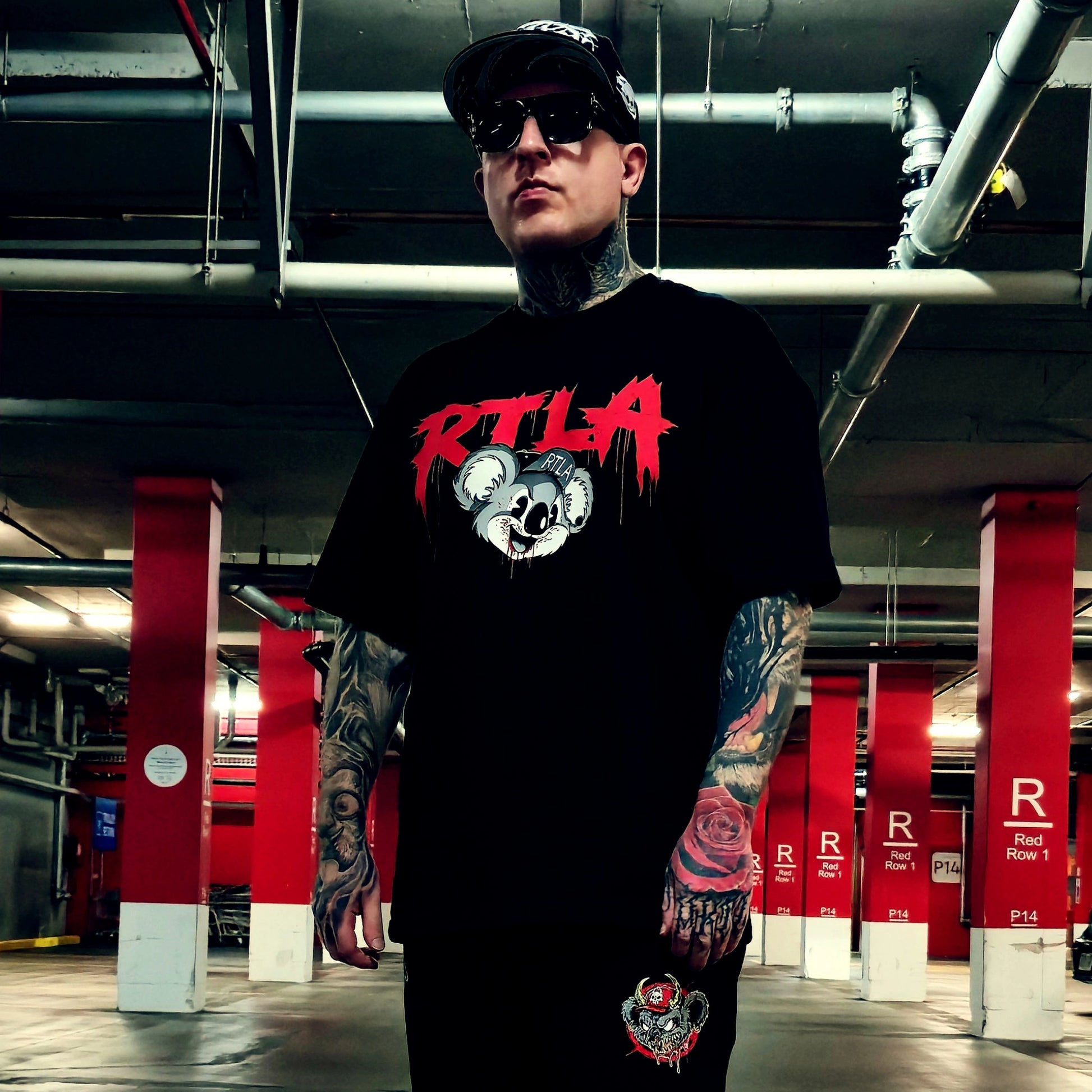 A tattooed man wearing a black cap, sunglasses, and a black t-shirt with a red and white 'RTL A' graphic stands in a parking garage. The background features red and white structural columns.
