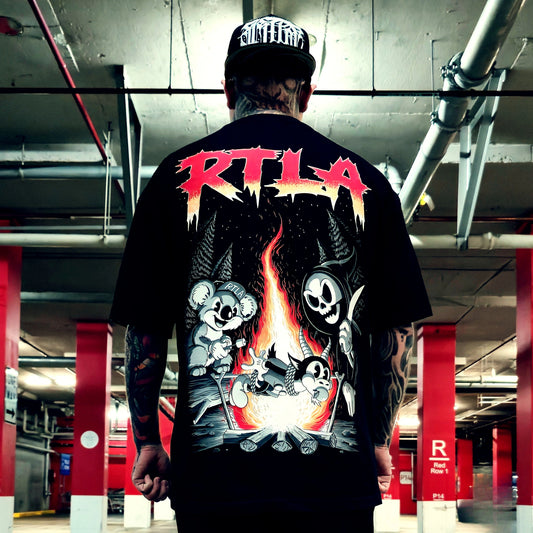 A man viewed from behind wearing a black graphic t-shirt and a black cap with a white logo, standing in a parking garage. The back of the t-shirt features a large graphic print with the text 'RTL A' in red and white, above cartoon koalas, a skeleton, and a campfire.