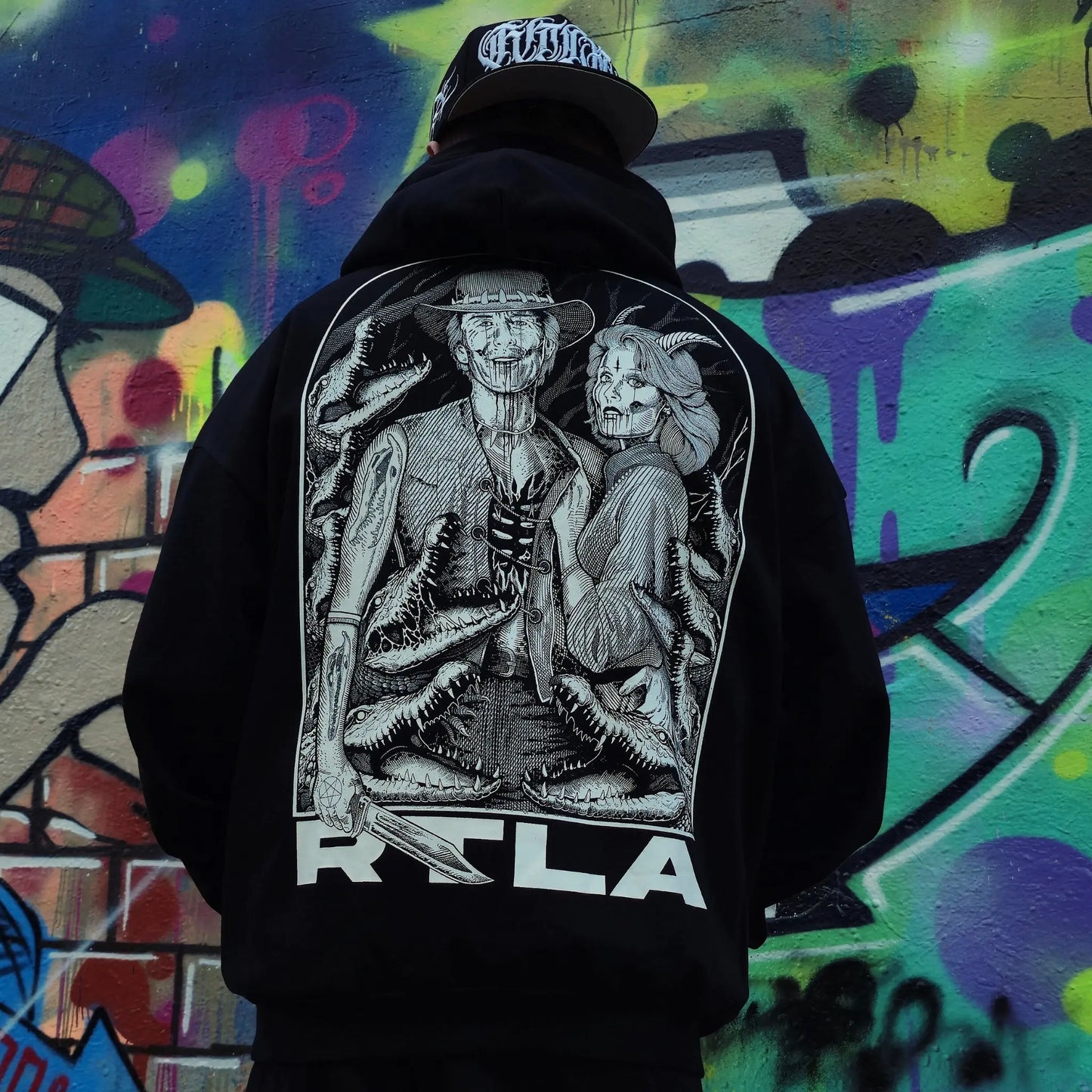 Person wearing a black hoodie with a graphic design on the back against a colourful graffiti background. RTLA-Apparel