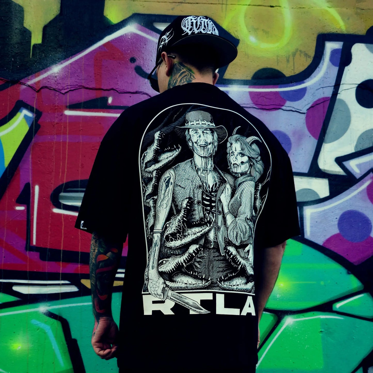 Person wearing a black t-shirt with a graphic design in front of colourful graffiti. RTLA-Apparel