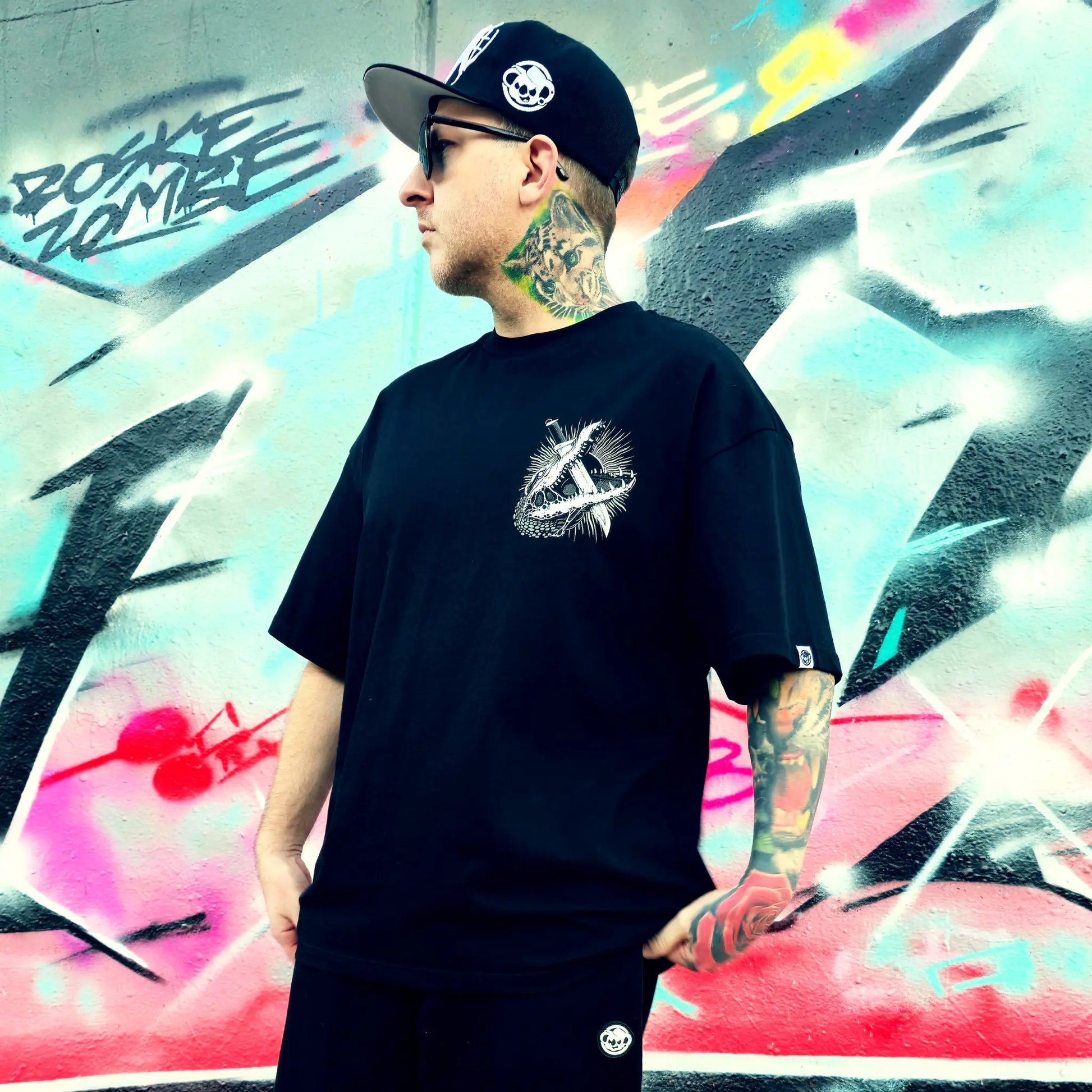 Person wearing a black t-shirt with a logo in front of a colourful graffiti wall. RTLA-Apparel