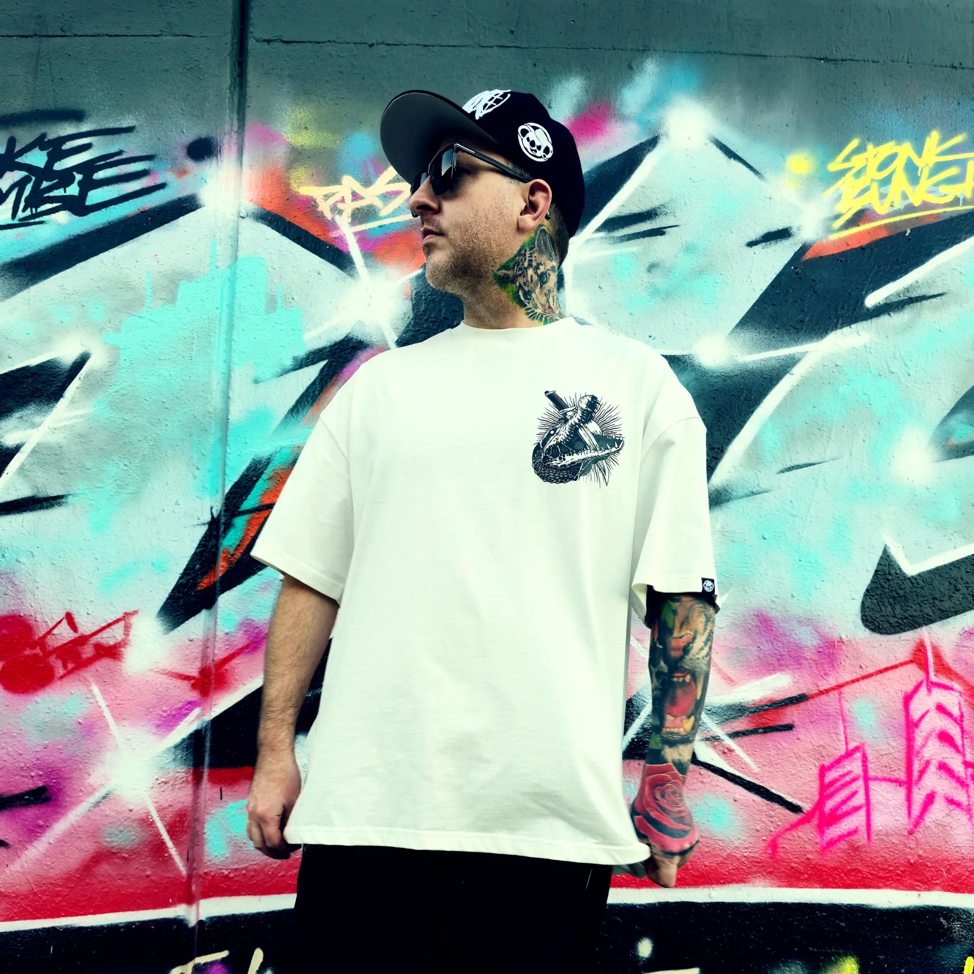 Man wearing a white t-shirt with a graphic design, black cap, and sunglasses in front of a colourful graffiti wall. RTLA-Apparel