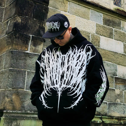 Person wearing a black hoodie with white designs and a cap, standing against a stone wall.