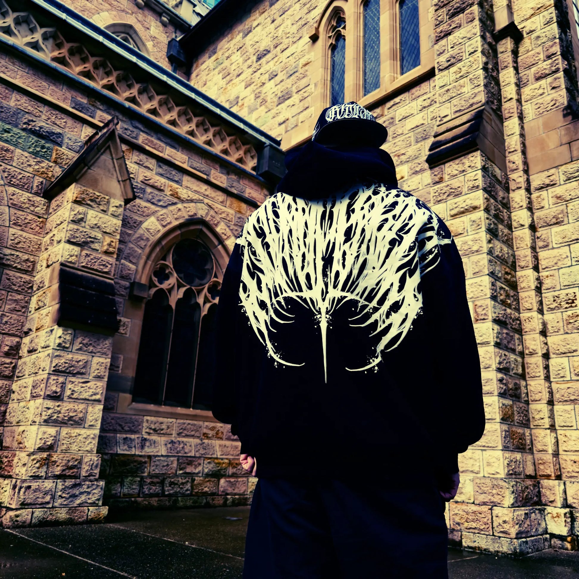 Person wearing a dark hoodie with a white pattern in front of a stone building