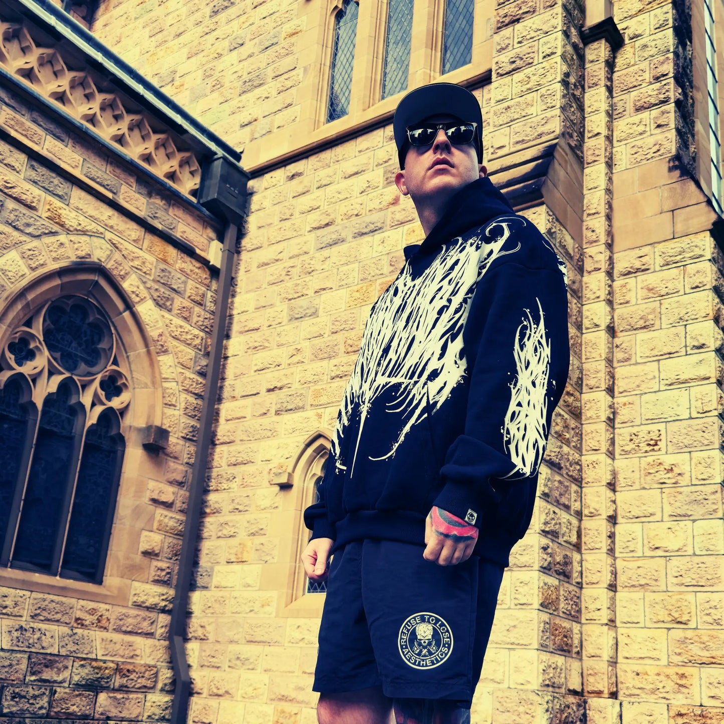 Person wearing a black and white jacket with flame design, sunglasses, and blue shorts with a logo, standing against a stone building.