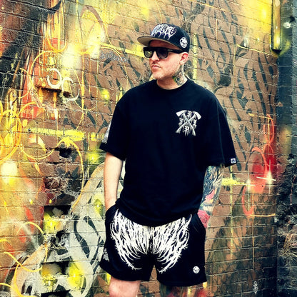 Person wearing a black t-shirt and shorts with a graphic design, standing in front of a graffiti-covered wall.