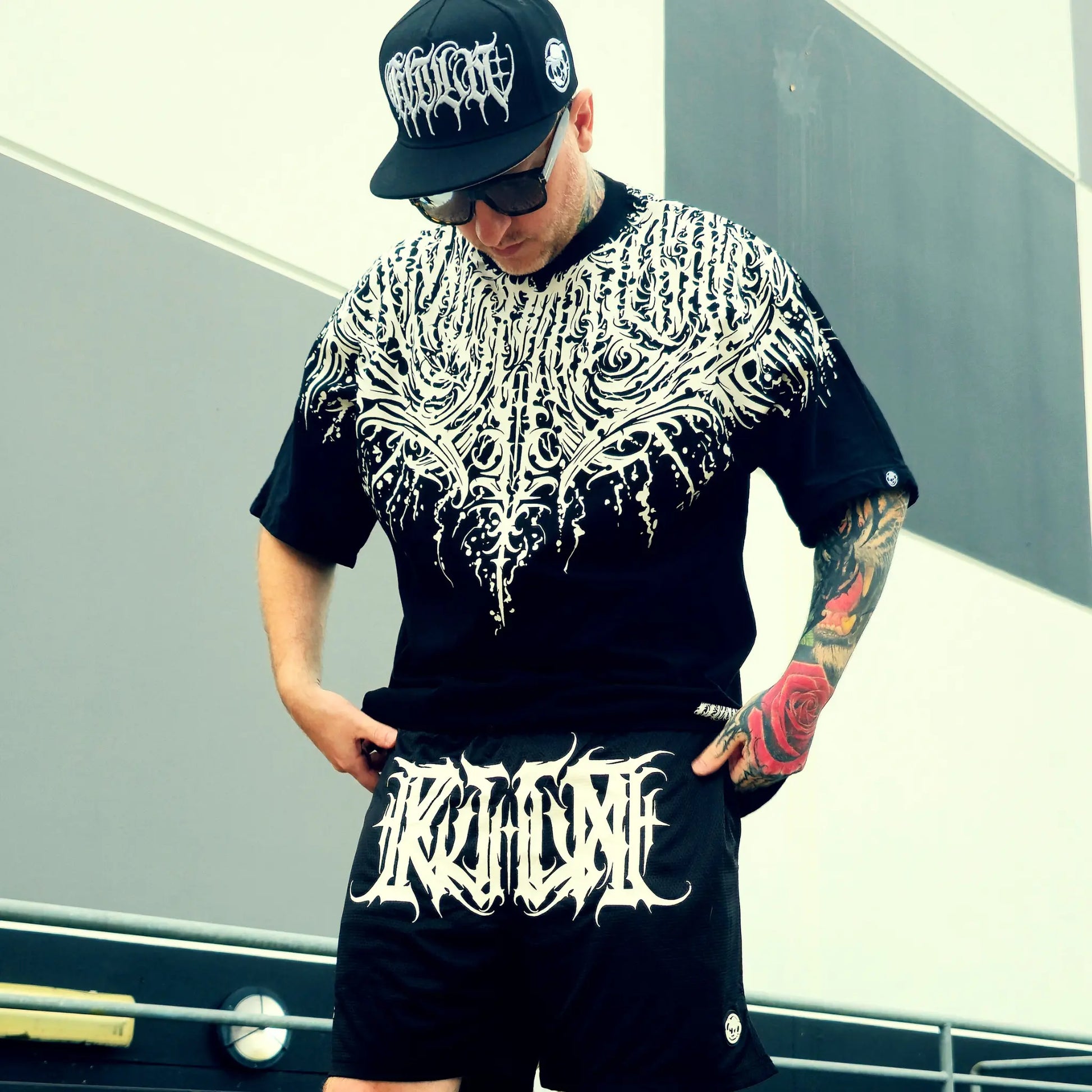 Person wearing a black and white patterned shirt and shorts with 'Kulture' branding, standing against a gray wall.