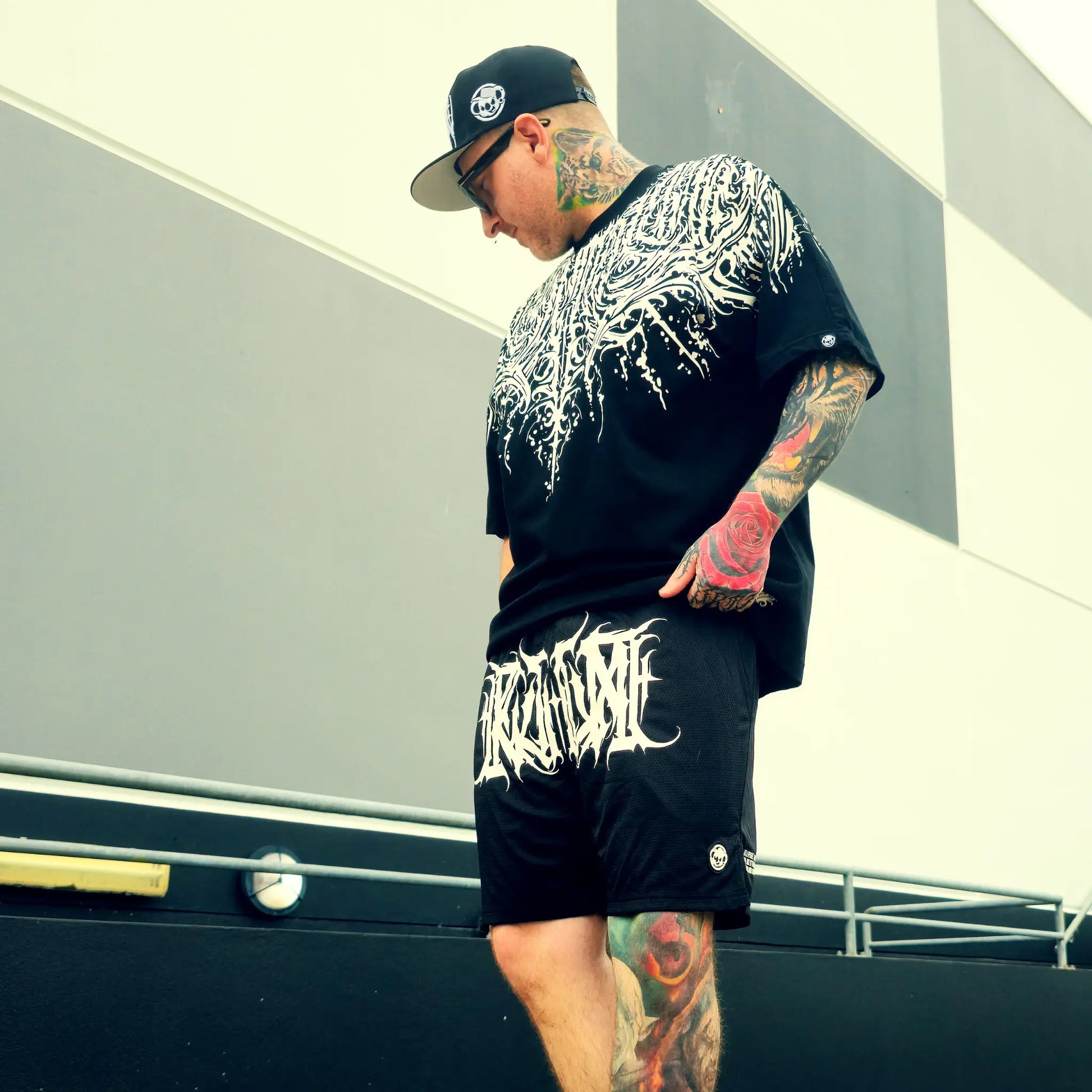 Man wearing a black graphic t-shirt and shorts with tattoos, standing outdoors.