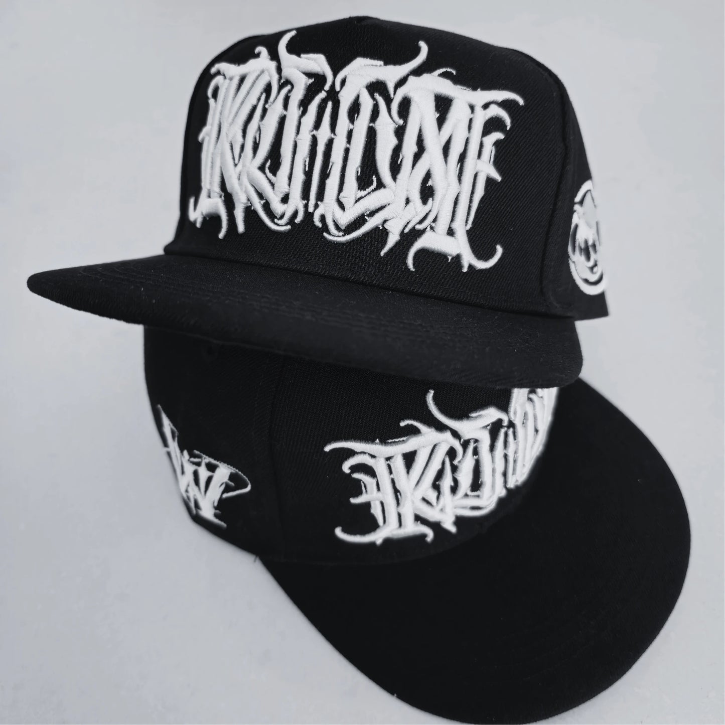 A black snapback cap with white 3D embroidered lettering on the front and a logo on the side. RTLA Apparel