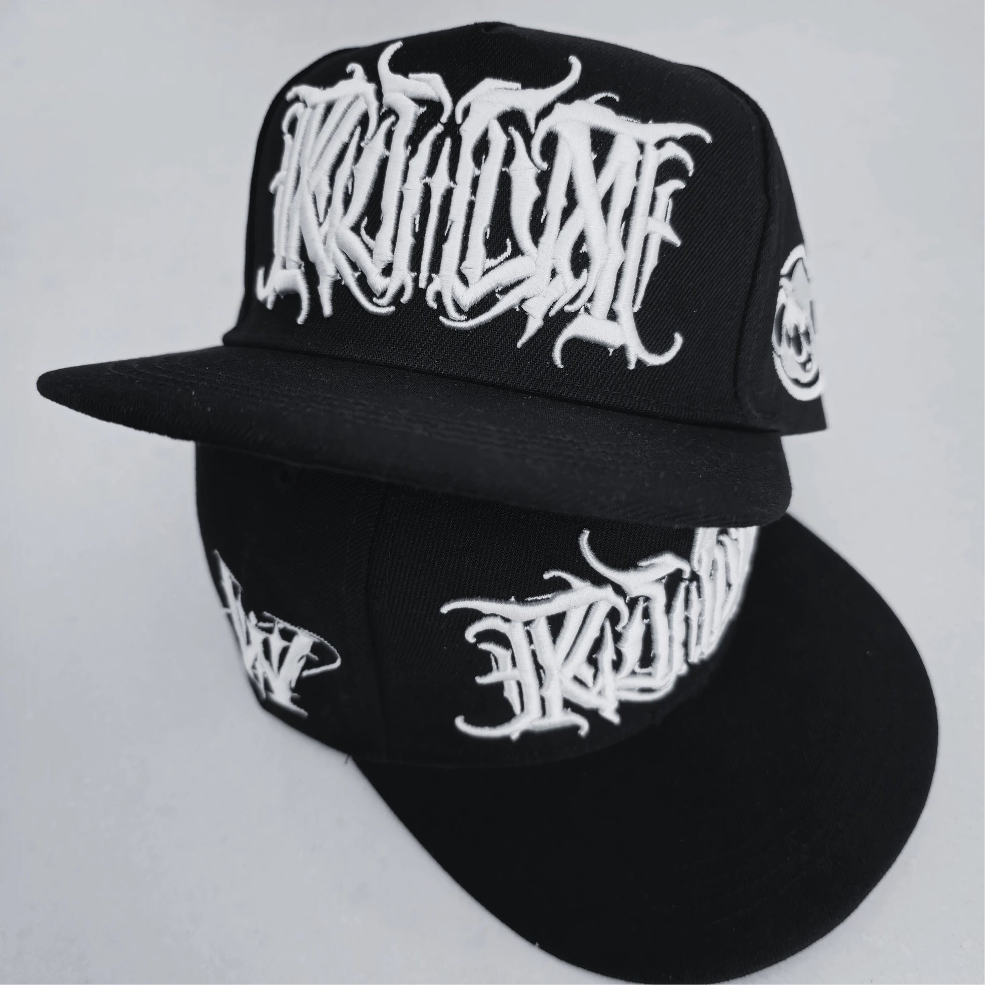 A black snapback cap with white 3D embroidered lettering on the front and a logo on the side. RTLA Apparel