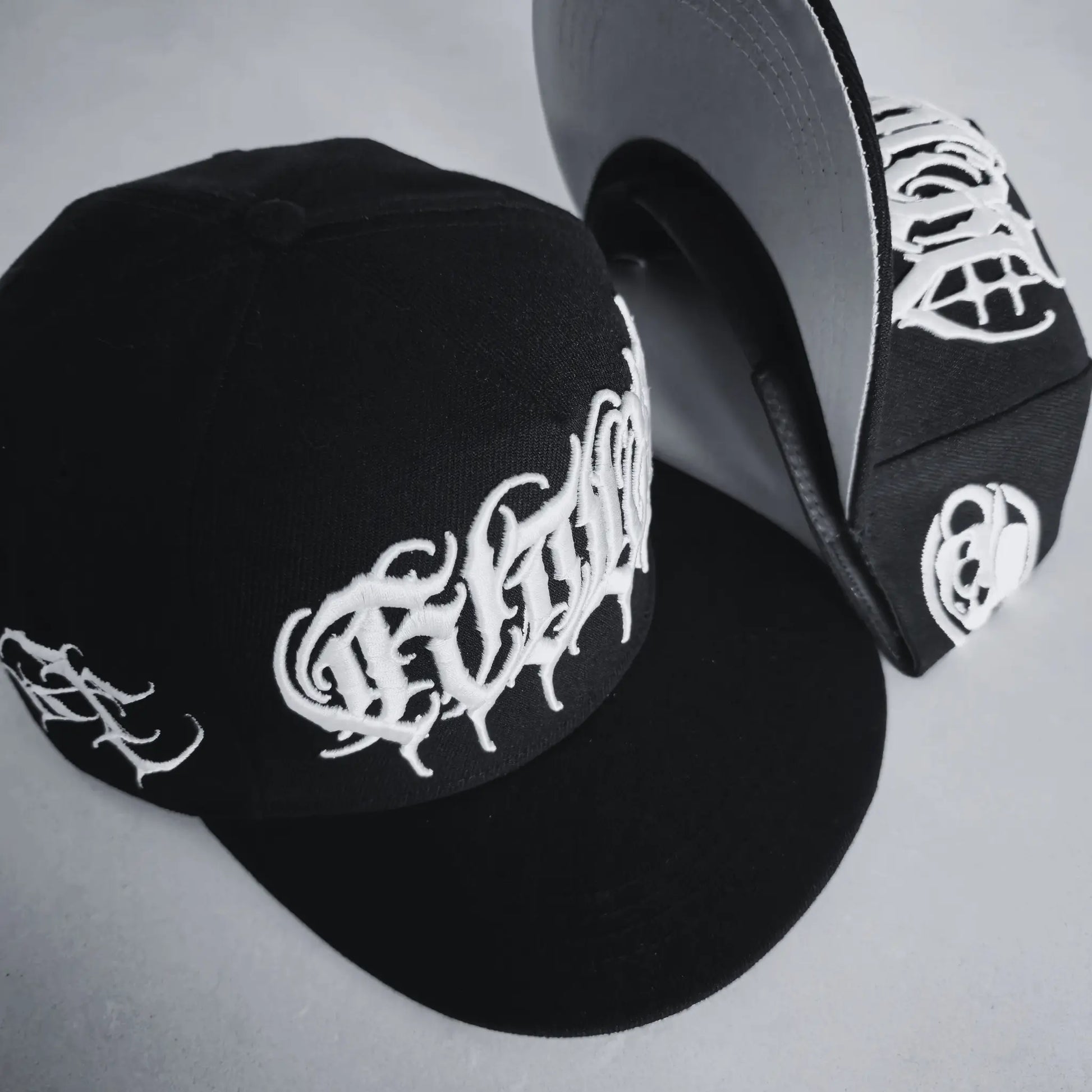 A black snapback cap with white 3D embroidered lettering and an artist signature logo on the side. RTLA-Apparel