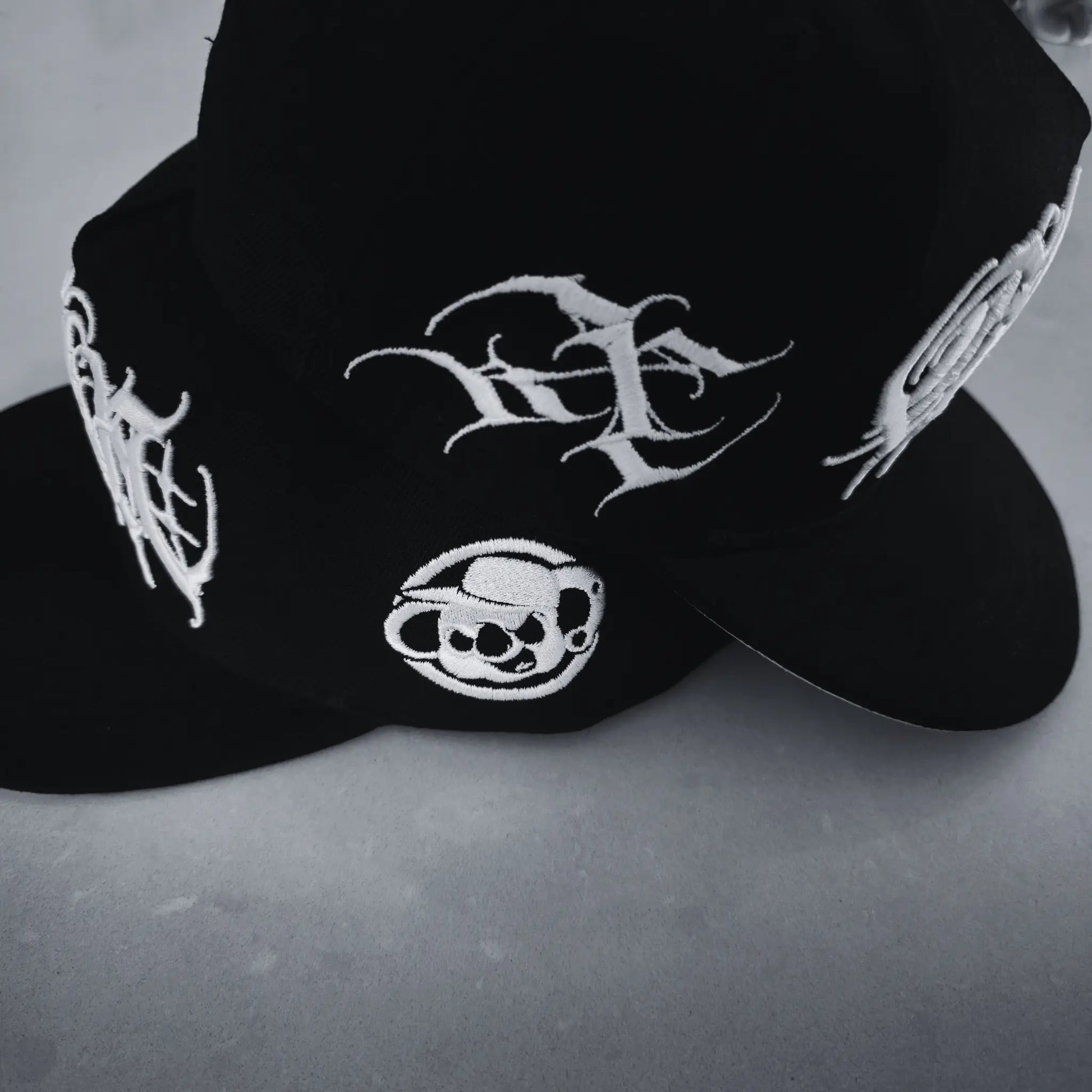 A black snapback cap with white 3D embroidered lettering and an artist signature logo on the side. RTLA-Apparel
