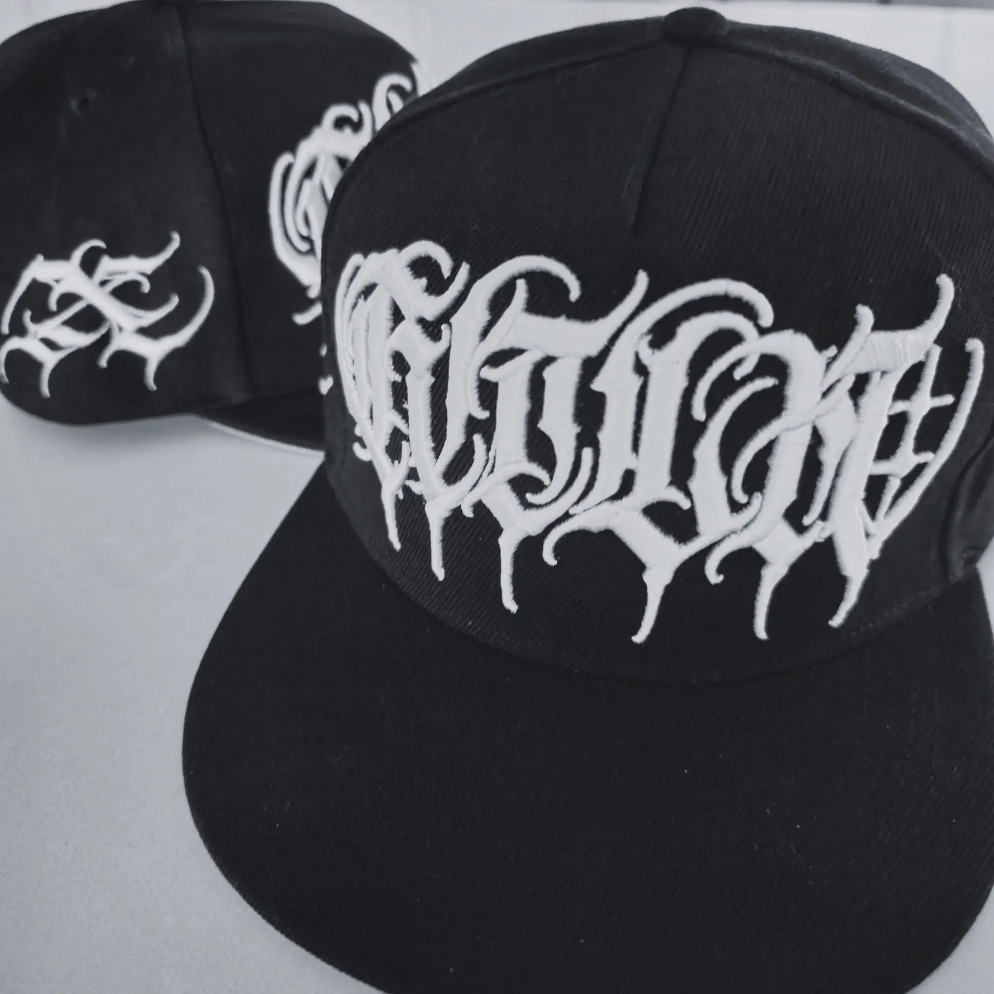 Two black snapback caps with white 3D embroidered lettering designed by Xover.Letters, featuring the artist's signature X logo. RTLA-Apparel