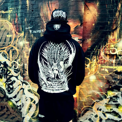 Person wearing a black hoodie with a white graphic design in front of a graffiti-covered wall.