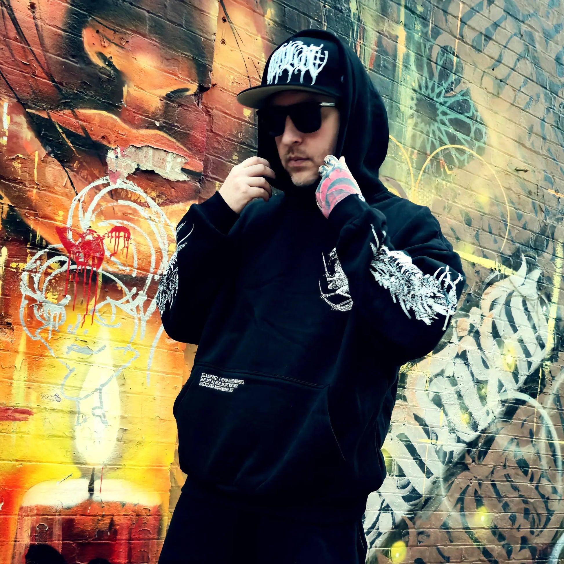 Person wearing a black hoodie and cap in front of a colorful graffiti wall.