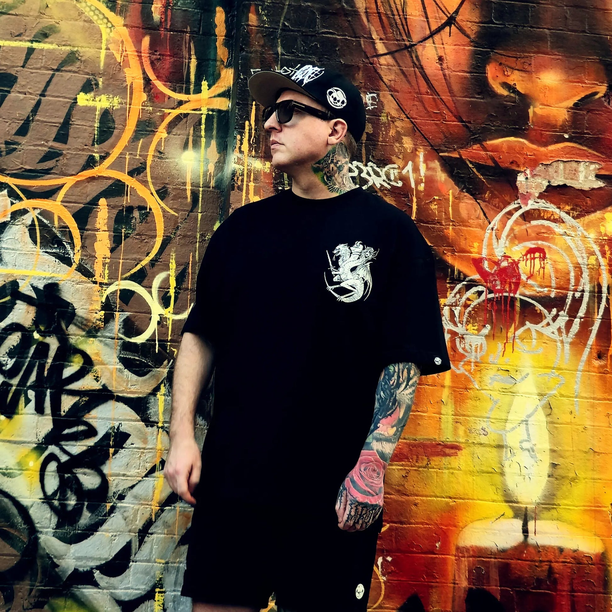 Person wearing a black t-shirt with a logo in front of a colorful graffiti wall.