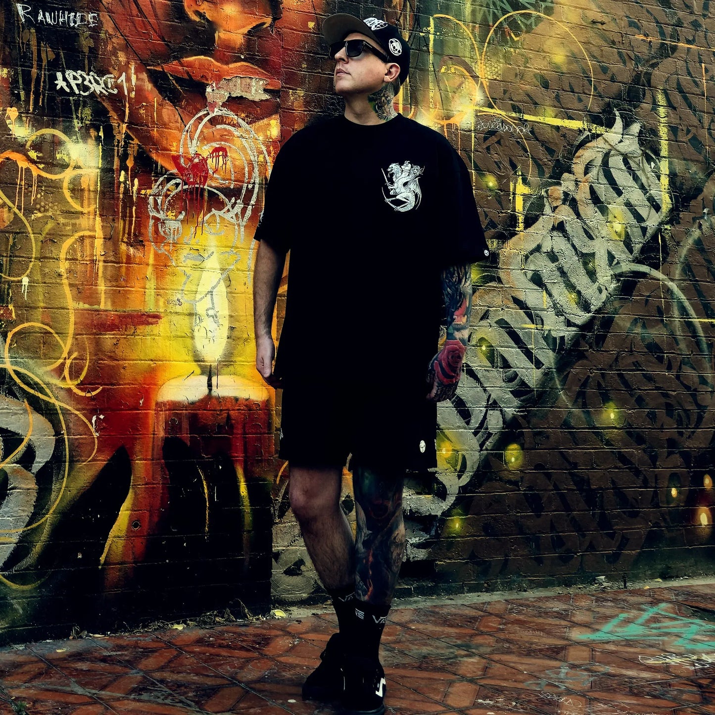 Person wearing a black outfit with a logo, standing in front of a graffiti-covered wall.