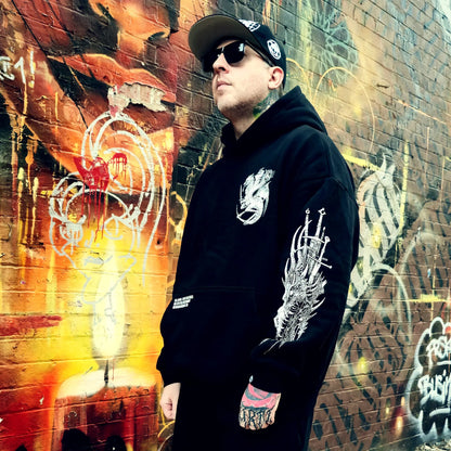 Person wearing a black hoodie with white designs in front of a graffiti-covered wall.