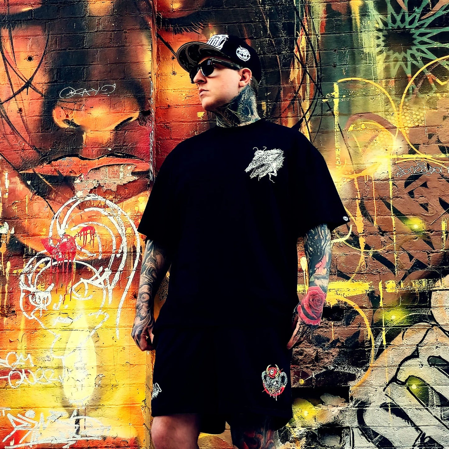 Person wearing a black t-shirt and shorts with tattoos standing in front of a colorful graffiti wall.