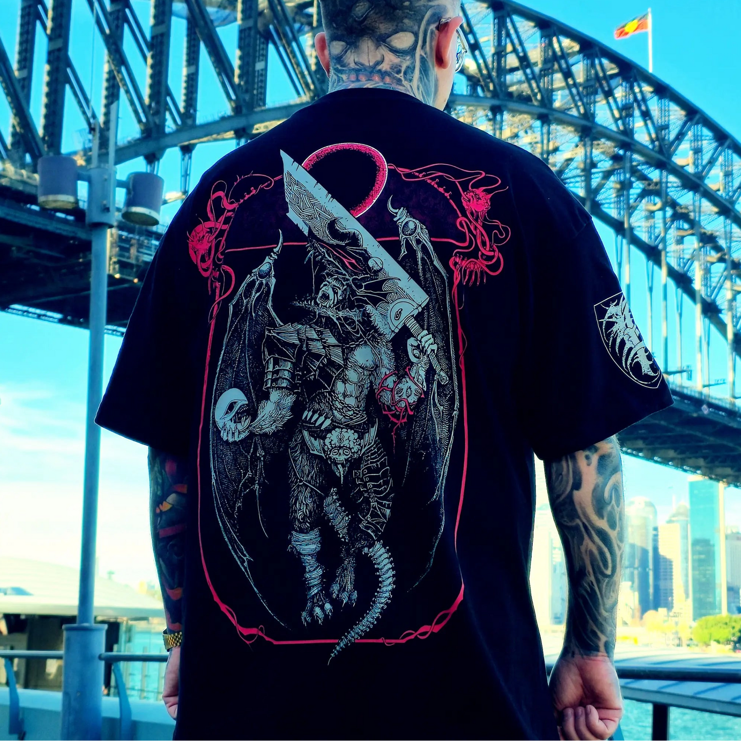 Person wearing a black t-shirt with a graphic design of a warrior and dragon, standing in front of a bridge.