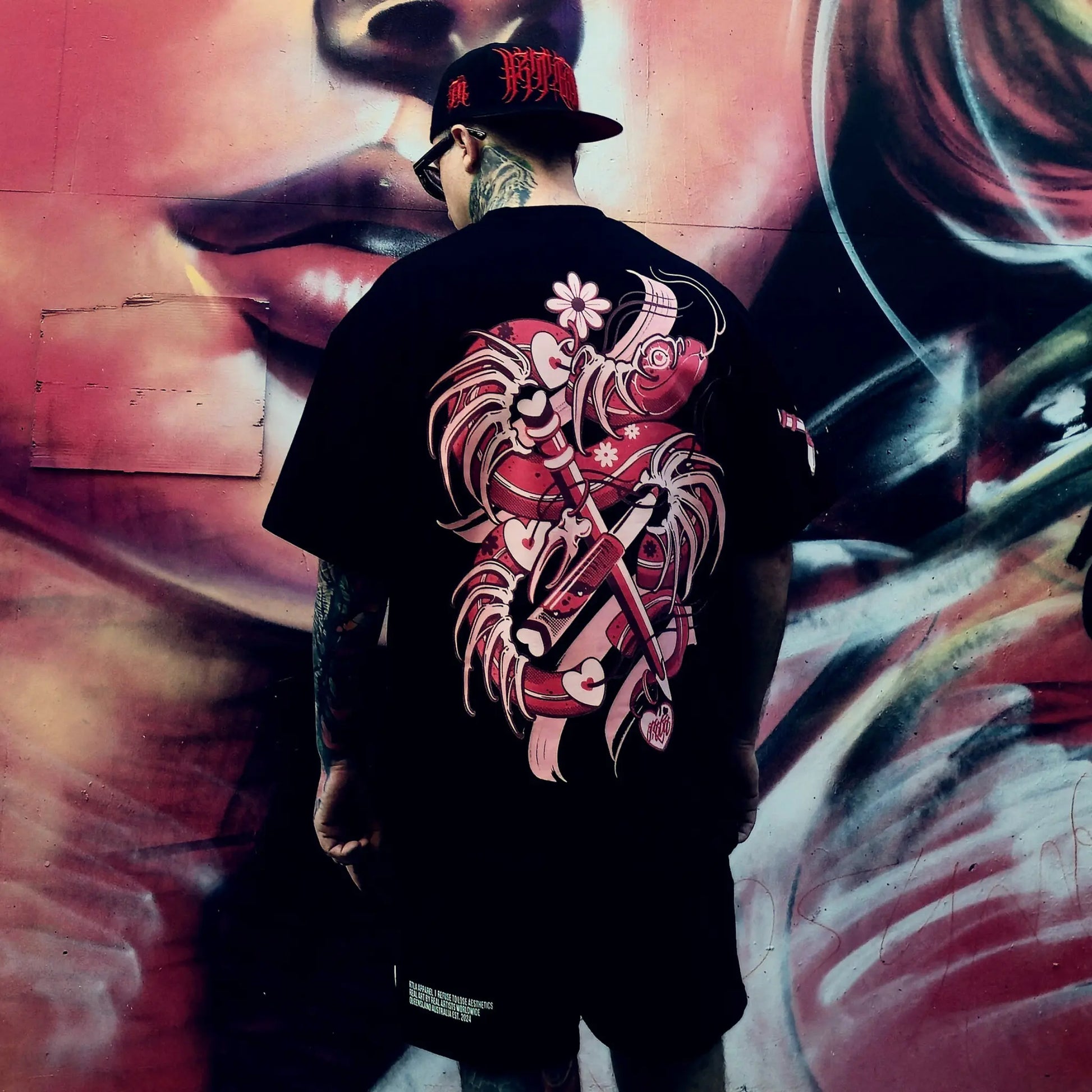 Person wearing a black t-shirt with a red and white graphic design, standing in front of a graffiti wall.